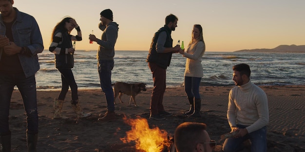 Photo heureux jeunes amis insouciants s'amusant et buvant de la bière par bonefire sur la plage alors que le soleil commence à se coucher