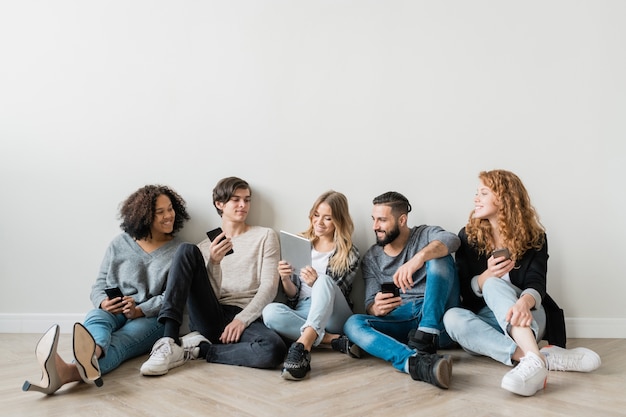 Heureux jeunes amis interculturels avec des gadgets assis en ligne par mur et regarder une vidéo drôle dans le pavé tactile tenu par une fille blonde