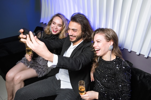 Heureux jeunes femmes glamour avec champagne et homme élégant avec smartphone faisant selfie sur canapé en boîte de nuit