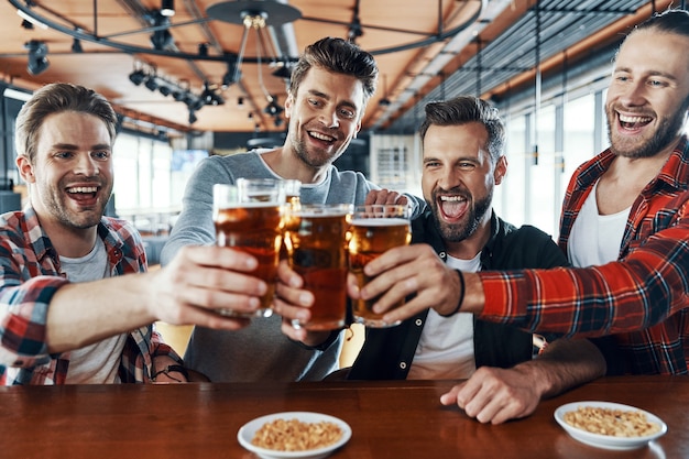 Heureux jeunes hommes en vêtements décontractés se grillant avec de la bière et riant assis dans le pub
