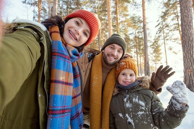 Heureux jeunes parents et leur mignonne petite fille en vêtements d'hiver chauds faisant du selfie contre l'environnement naturel par une journée d'hiver glaciale