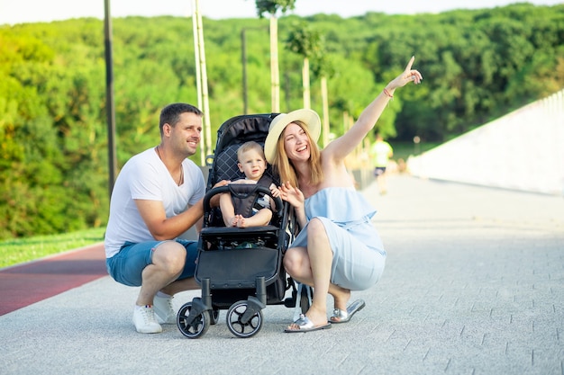 Heureux jeunes parents maman et papa marchent avec un bébé dans une poussette dans le parc en été au soleil couchant et sourient tout en s'amusant à pointer au loin