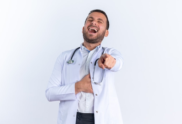 Heureux et joyeux jeune homme médecin en blouse blanche et avec un stéthoscope autour du cou en riant en pointant l'index vers vous debout sur un mur blanc