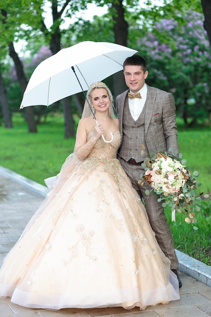 Heureux mariés avec un parapluie blanc sous la pluie, en été dans le parc. Mariage en plein air.