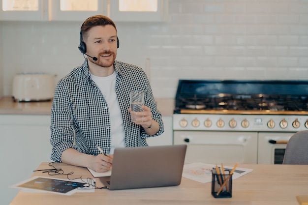 Heureux mec barbu dans des vêtements décontractés et un casque travaillant à distance sur un ordinateur portable à la maison, tenant un verre d'eau et prenant des notes tout en étudiant en ligne. Concept de travail à distance et d'enseignement à distance