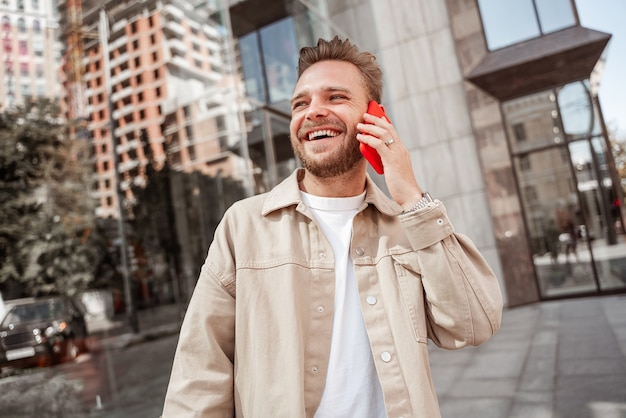 Heureux mec blond hipster lors d'une conversation sur smartphone avec le meilleur ami debout sur des étudiants de fond de rue urbaine communiquant via un gadget de téléphone portable, skateur millénaire