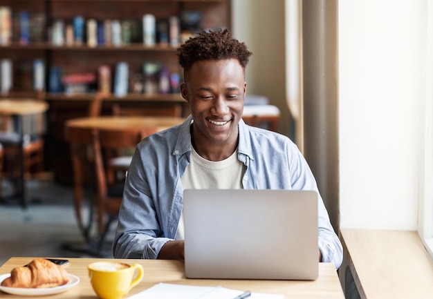 Heureux mec noir travaillant en ligne sur un ordinateur portable au café