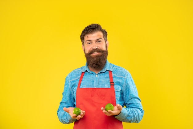 Heureux mec en tablier tenant des citrons verts frais agrumes fond jaune, marchand de légumes.