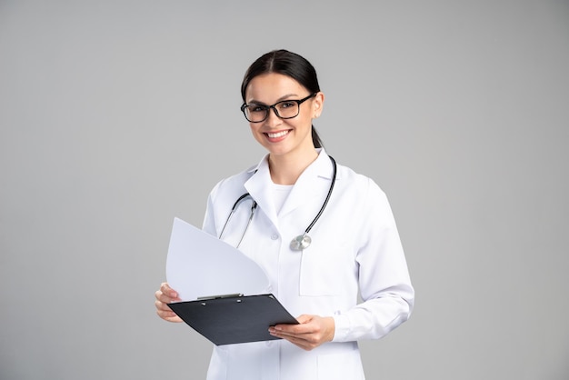 Heureux médecin caucasien avec stéthoscope tenant le presse-papiers et lisant quelque chose en se tenant debout et souriant isolé sur fond gris