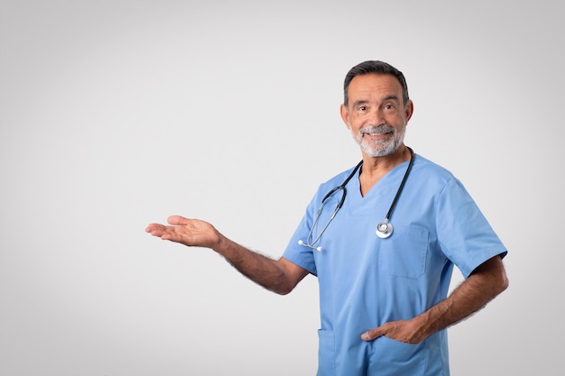 Heureux médecin senior caucasien en uniforme bleu tenir un espace vide à portée de main isolé sur fond gris