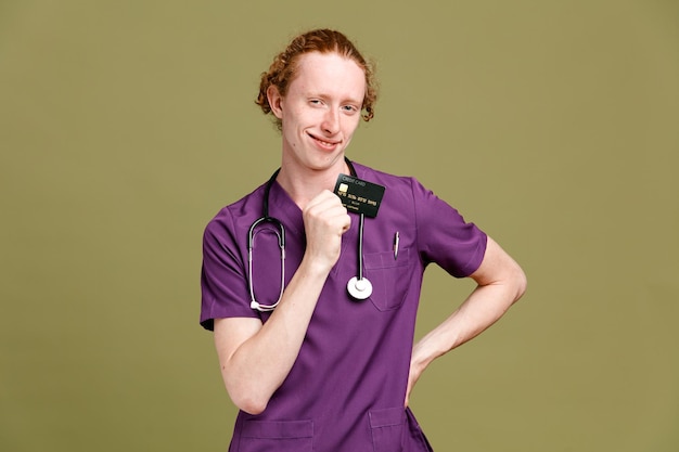 Heureux de mettre la main sur les hanches jeune homme médecin portant l'uniforme avec stéthoscope tenant la carte isolé sur fond vert