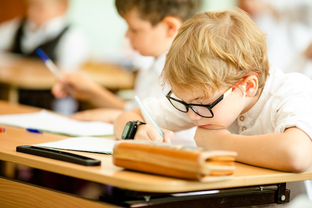 Heureux mignon garçon intelligent est assis à un bureau dans un verre avec une main levée. Retour à l'école