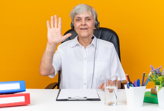 Heureux opérateur de centre d'appels féminins de race blanche sur un casque assis au bureau avec des outils de bureau levant la main