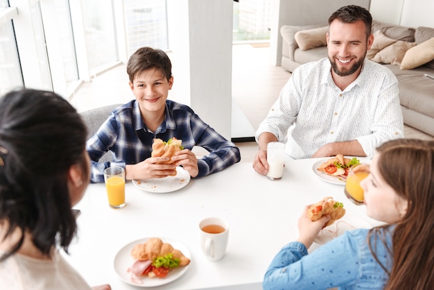 heureux parents avec enfants 8-10, assis à table dans une cuisine lumineuse et prendre le petit déjeuner tout en mangeant des sandwichs au croissant