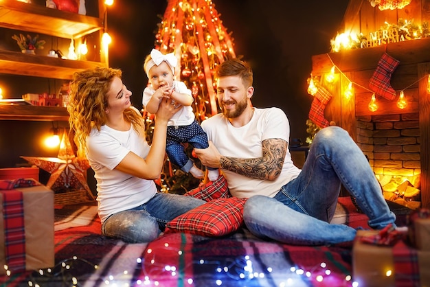 Heureux parents jouant avec leur petite fille assis ensemble près de l'arbre de Noël et de la cheminée