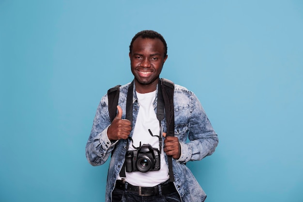 Heureux passionné de photographie avec un appareil photo reflex numérique donnant le symbole de la main d'approbation. Jeune photographe montrant le geste de la main tout en ayant un appareil photo professionnel sur fond bleu.