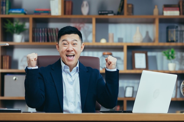 Heureux patron asiatique regarde la caméra et célèbre la victoire de l'entreprise tient les mains levées geste de triomphe