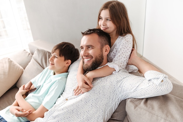 Heureux père de 30 ans avec fils et fille étreignant ensemble sur le canapé à la maison, et regardant de côté