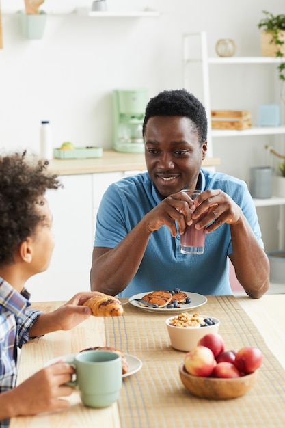 Heureux père africain prenant son petit déjeuner avec son fils, ils sont assis à la table et parlent dans la cuisine