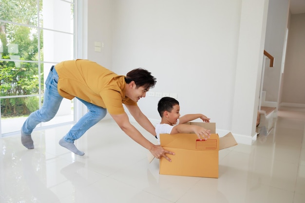 Heureux père asiatique jouant avec de mignons petits enfants fils riant le jour du déménagement garçon s'amusant à rouler dans une boîte dans une nouvelle maison