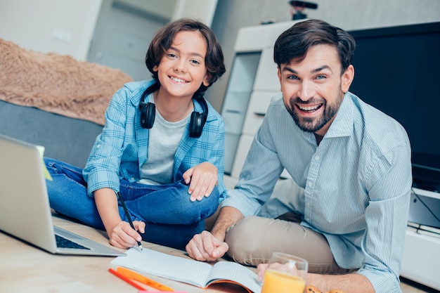 Heureux père faisant ses devoirs avec son fils sur le sol devant un ordinateur portable Cahier d'exercices et crayon dans les mains