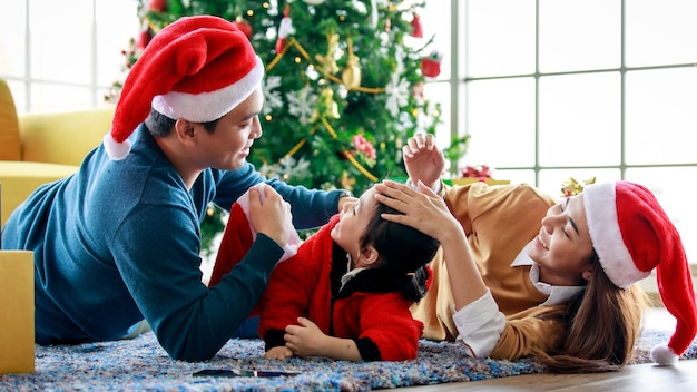 Heureux père de famille asiatique, la mère et la fille portent un pull avec un chapeau de père Noël rouge et blanc allongé sur un tapis célébrant le réveillon de Noël ensemble devant le décor du sapin de Noël.