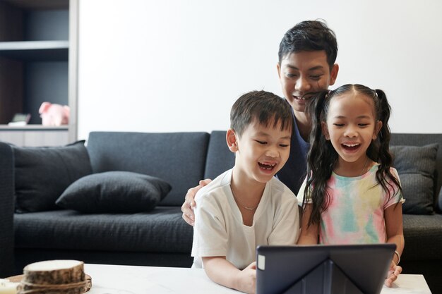 Heureux père riant, son fils et sa fille regardant un dessin animé drôle ou une émission sur une tablette numérique