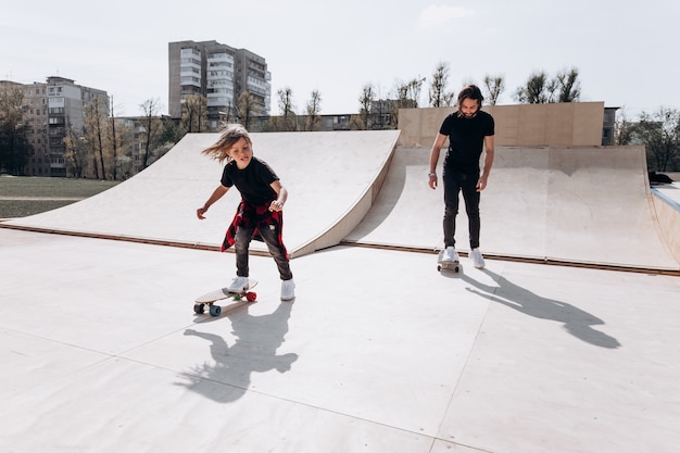 Heureux père et son petit fils vêtus de vêtements décontractés font du skateboard dans un skate park avec des toboggans à la journée ensoleillée.