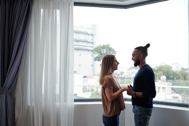 Heureux petit ami et petite amie se tenant la main et se regardant en se tenant contre la grande fenêtre de l'appartement