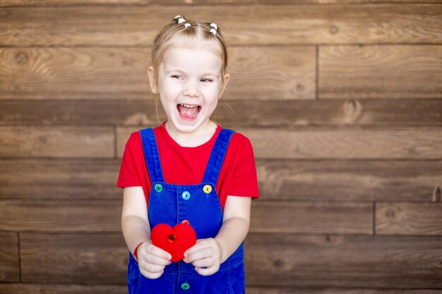 Heureux petit enfant une fille avec un petit coeur rouge dans ses mains sourit ou rit contre un mur en bois concept de la saint-valentin espace vide pour le texte