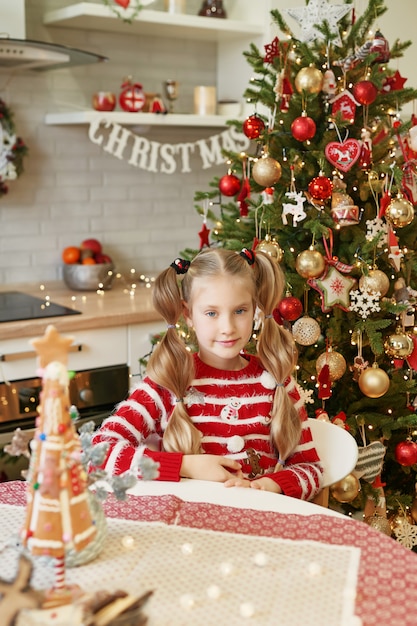 Heureux petit enfant, jolie fille enfant, assis à table dans la cuisine domestique. Cuisine avec arbre de Noël aux couleurs traditionnelles. Teen girl s'amuser à la maison.