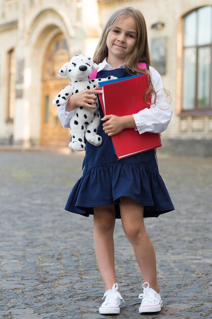Heureux petit enfant en uniforme scolaire tenant des livres d'étude et un chien jouet à l'extérieur le 1er septembre