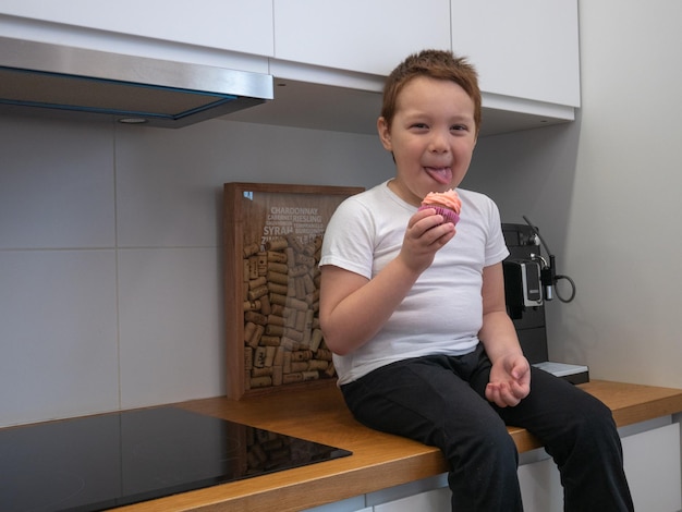 Heureux petit garçon asiatique assis sur une table de cuisine léchant la crème de cupcake souriant