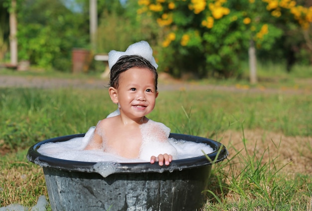 Heureux petit garçon asiatique prendre un bain avec bulle de mousse dans un bassin en plastique noir dans la nature