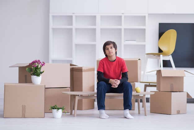 heureux petit garçon assis sur la table avec des boîtes en carton autour de lui dans une nouvelle maison moderne