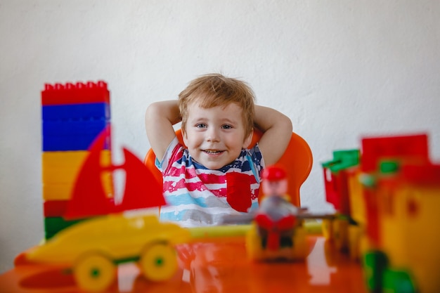 Un heureux petit garçon riant est assis à sa table parmi les jouets colorés avec ses mains en l'air. Photo de haute qualité