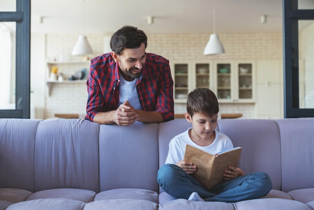 Heureux petit garçon et son père joyeux lisent un livre assis sur le canapé à la maison dans le salon.