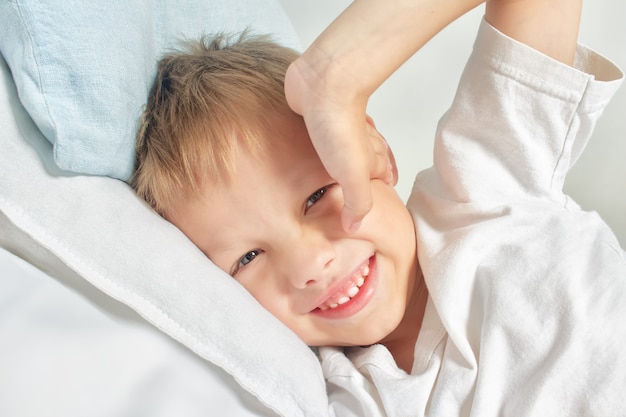 Heureux petit garçon souriant après s'être réveillé s'étendant dans son lit. Bonjour . Portrait d'un enfant blanc aux yeux gris et aux cheveux blonds, profitant de la vie à la maison.