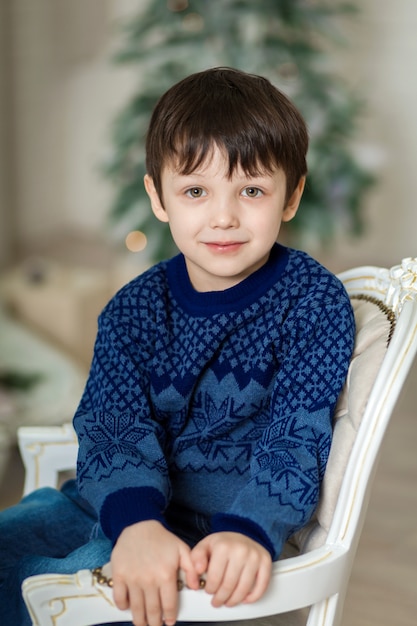 Photo heureux petit garçon souriant assis sur un fauteuil près de sapin de noël et tenant une boîte de cadeau de noël.