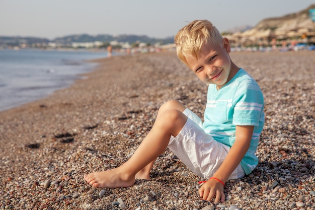 Heureux petit garçon souriant assis sur la plage et jouant avec des pierres