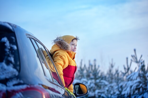 Heureux petit garçon souriant regarde d'une fenêtre de voiture par une journée ensoleillée dans la forêt enneigée d'hiver.