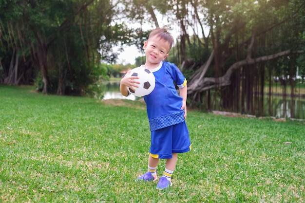 Heureux petit joueur de football en uniforme de football tient un ballon de football