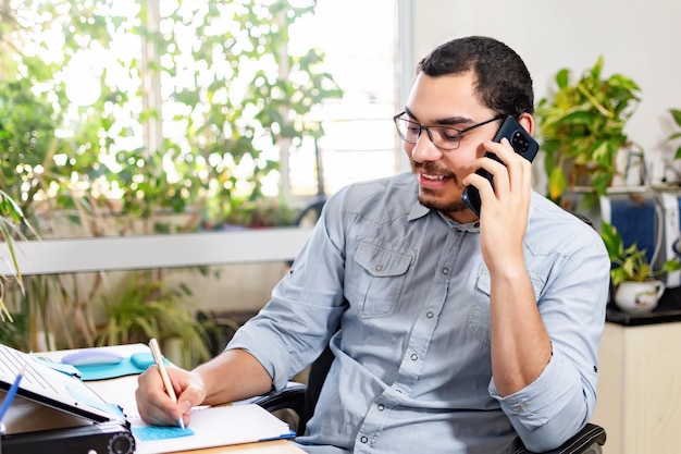 Heureux professionnel parlant sur un téléphone intelligent tout en écrivant dans un journal au bureau