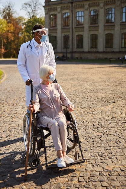 heureux professionnel de la santé déplaçant un fauteuil roulant avec un retraité, tenant un bâton