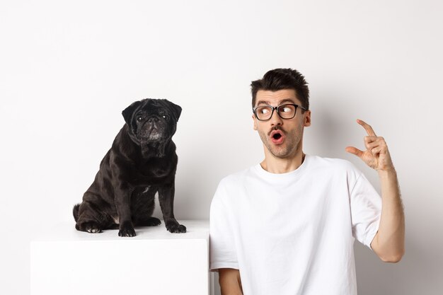 Heureux propriétaire de chien mâle assis près d'un mignon carlin noir, regardant un animal de compagnie avec un visage étonné et surpris, fond blanc