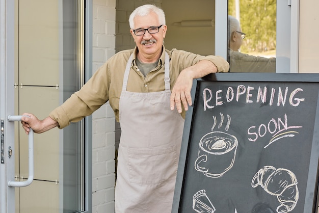 Heureux propriétaire mature de boulangerie debout près de l'entrée et de la porte d'ouverture