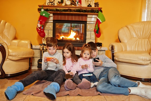 Heureux quatre enfants regardant dans leur téléphone portable à la maison près d'une cheminée dans un salon chaleureux le jour de l'hiver