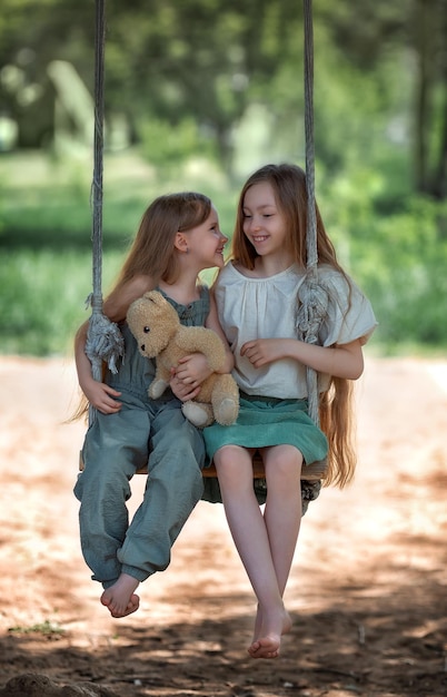 Heureux rire enfants filles sœurs aux cheveux longs bénéficiant d'une balançoire avec un ours en peluche sur une journée d'été ensoleillée