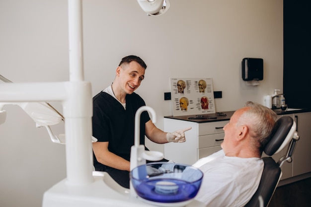 Heureux et souriant homme âgé de 7075 ans avec des dents blanches lors de l'examen d'un dentiste assis dans une chaise clinique dentaire À la recherche d'un jeune médecin dentiste Soins dentaires pour les personnes âgées Concept de soins de santé