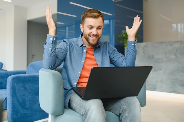 Heureux souriant homme de travail en ligne à distance en tenue décontractée avec ordinateur portable dans un geste gagnant joyeux et réussi assis dans un bureau de coworking à un bureau